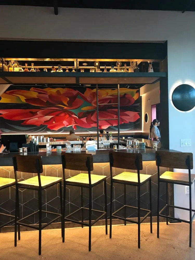 Modern bar with a black countertop, five bar stools with yellow seats, and a colorful floral mural on the back wall. Glassware and bar essentials are on the counter, and the bar features a mezzanine with bottles on display.