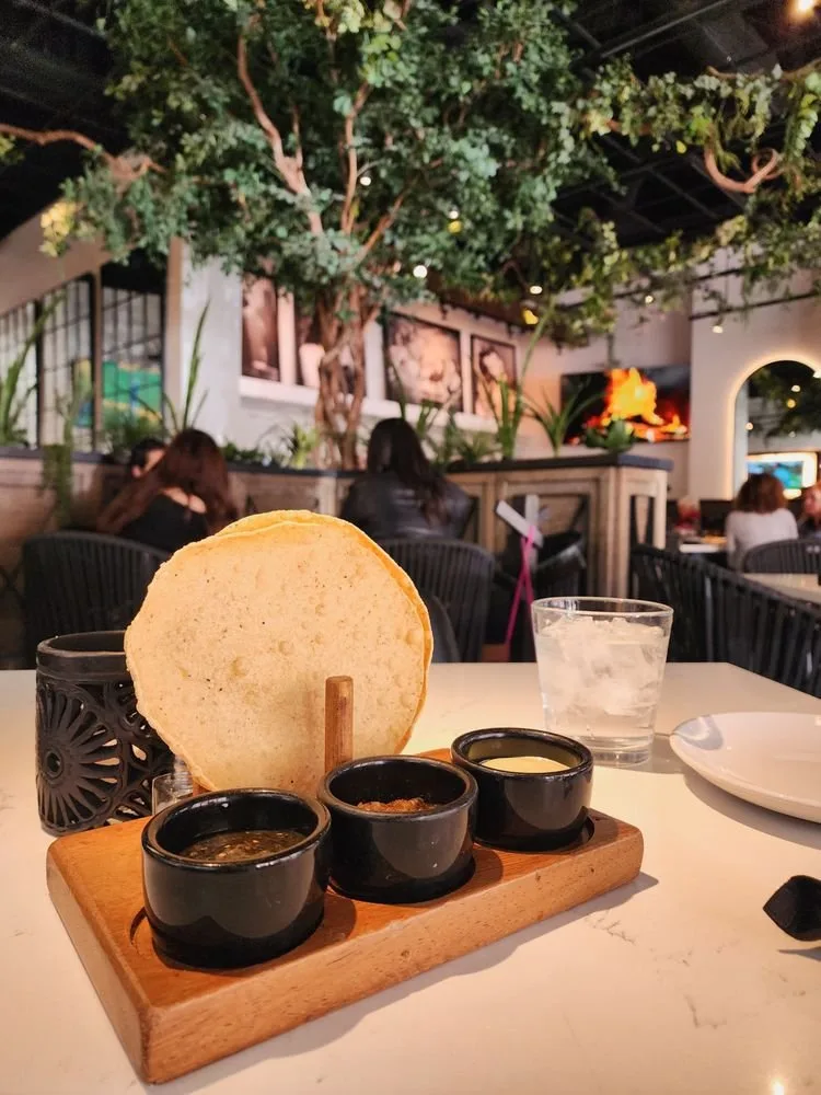 A small plate with three black bowls of different sauces, a large round bread, a glass of water, and a white plate on a white table in a restaurant with indoor trees and people dining in the background.