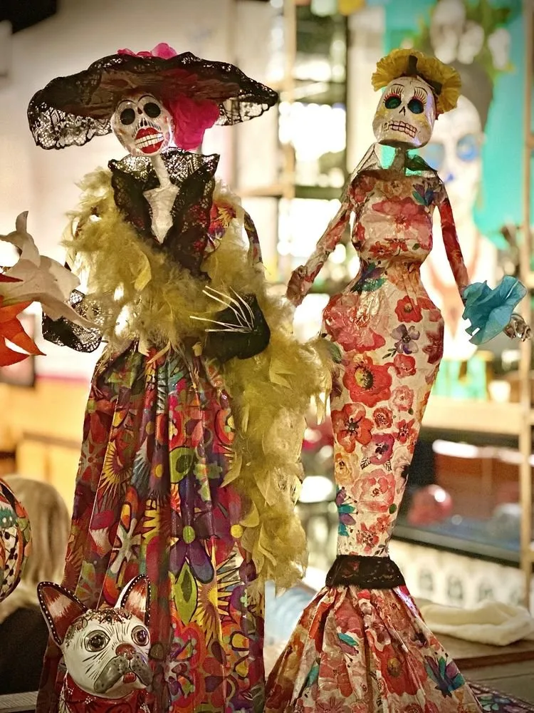 Decorative skeleton and doll figures dressed in colorful floral and patterned fabrics, with skeletal faces, set in an indoor environment.