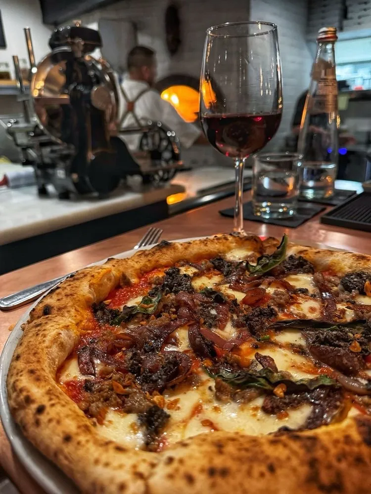 A close-up of a pizza with cheese, meat, and herbs on a wooden table, with a glass of red wine, a water bottle, and a fork in the foreground. The background shows a bar counter and a person working behind it.