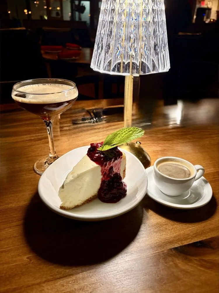 A slice of cheesecake with berry topping and a mint leaf on a white plate, a small cup of coffee, and a cocktail on a wooden table in a dimly lit setting with a decorative glass lamp.