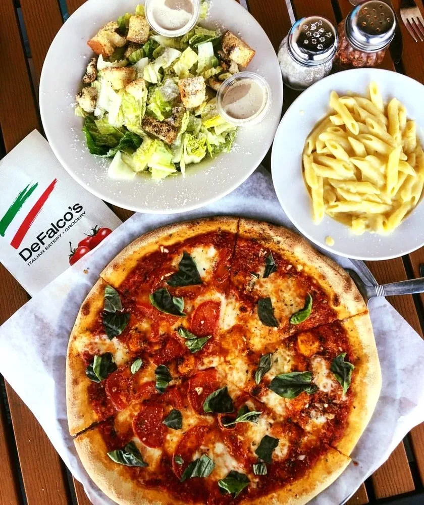 A pizza with tomato slices, melted cheese, and basil leaves, a side salad with croutons and dressing, a plate of cooked pasta, and salt and pepper shakers on a wooden table at DeFALCO'S Italian Eatery and Grocery.