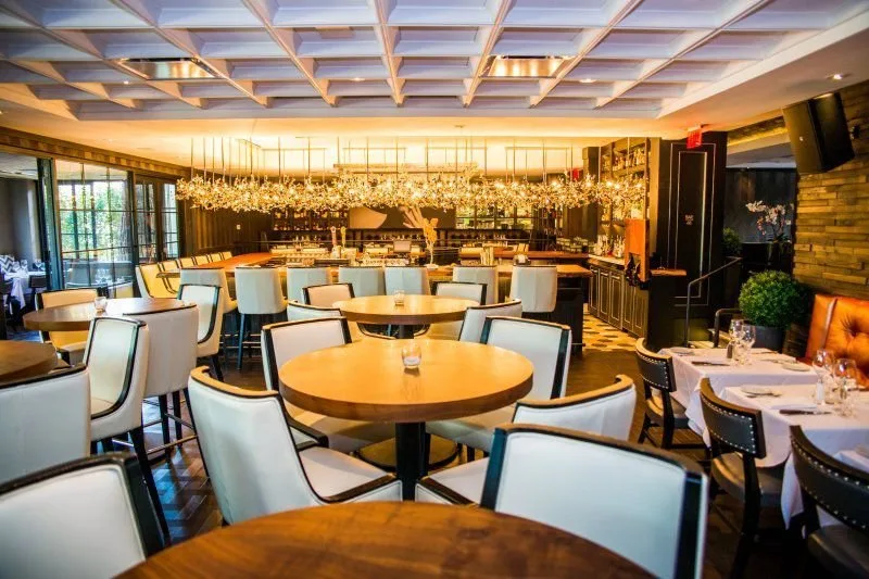 Empty upscale restaurant with round and rectangular tables, white and beige chairs, modern decor, large windows, and decorative lighting fixtures.