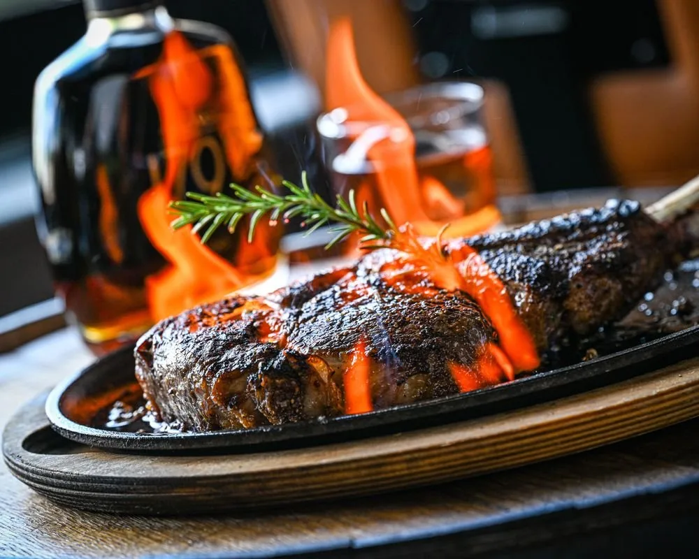 A grilled steak on a sizzling platter with a sprig of rosemary, with flames visible in the background and a glass of whiskey.