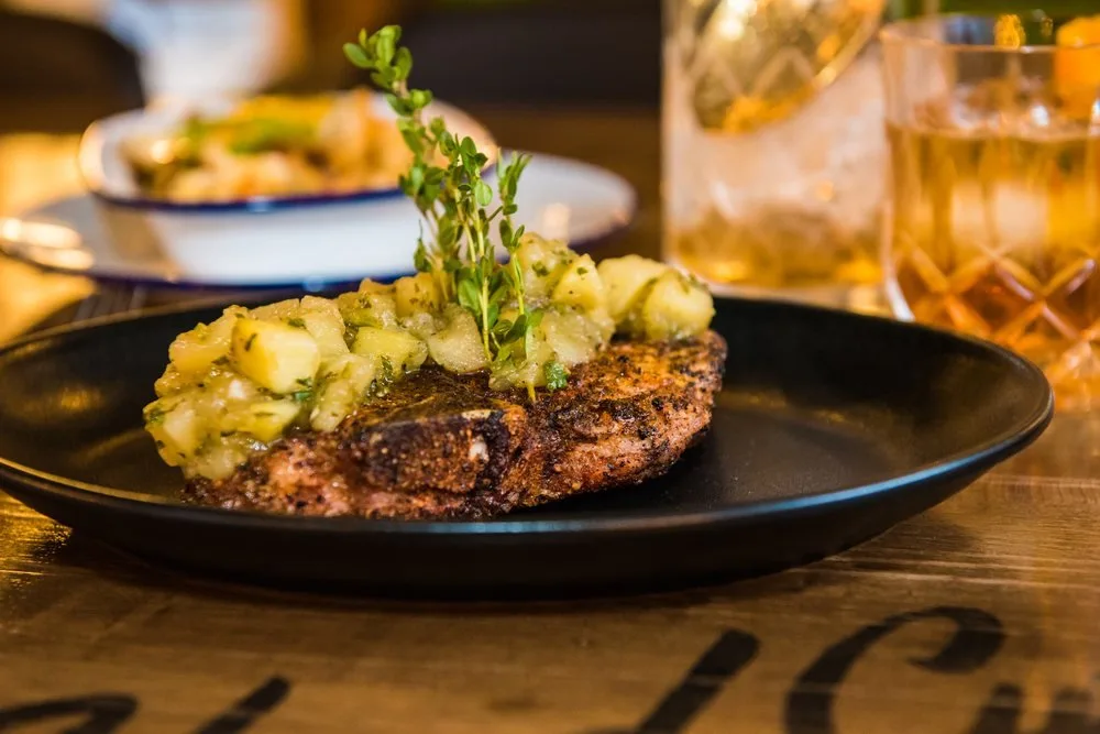 Grilled steak topped with a green herb and vegetable relish, garnished with fresh herbs, served on a black plate with drinks in the background.