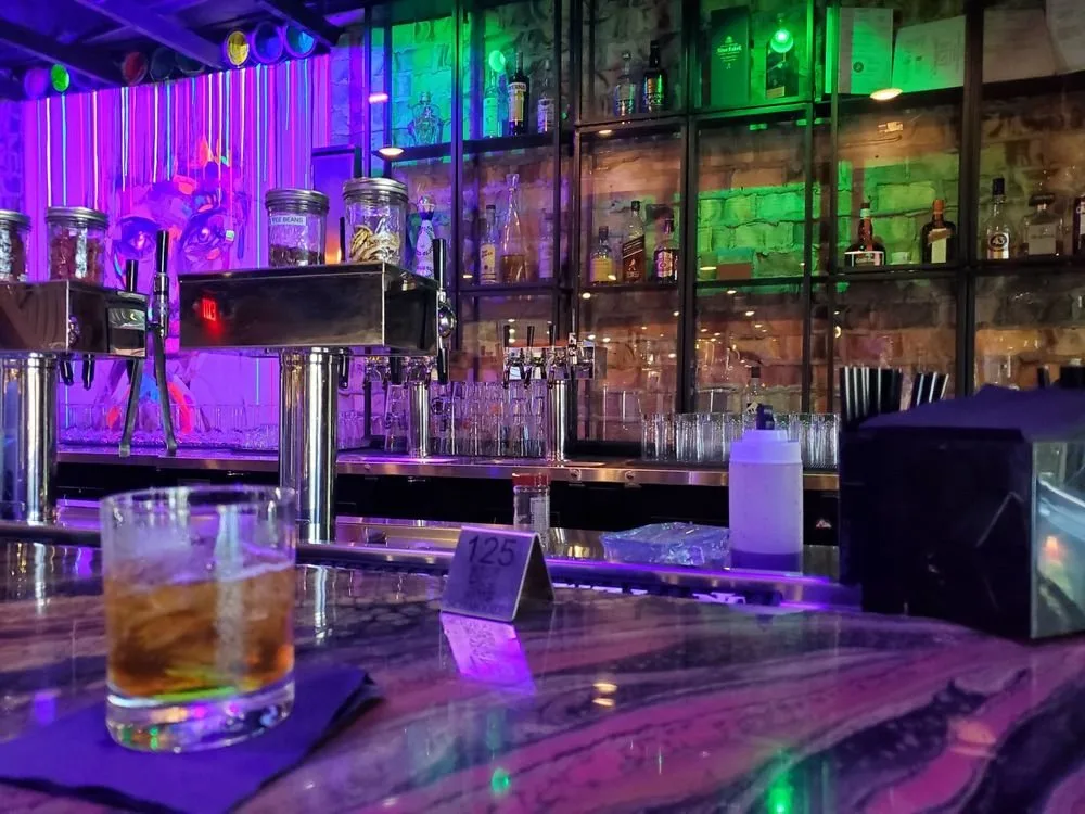 Bar counter with a glass of whiskey on a napkin, colorful neon lights, liquor bottles on shelves, and a row of glasses.