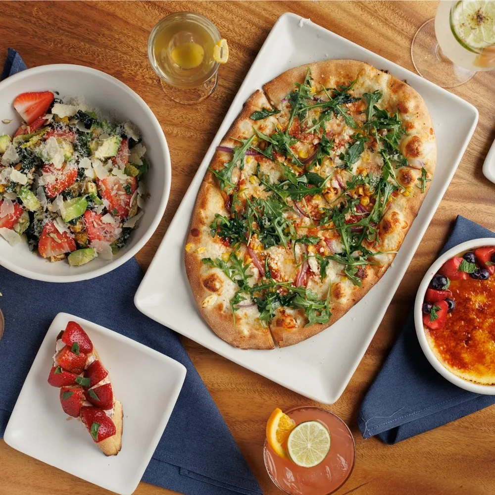 A table with a large pizza topped with arugula, a bowl of strawberry salad with parmesan cheese, a slice of strawberry tart, a glass of lemon and ice drink, and a small glass of champagne with lemon slice.