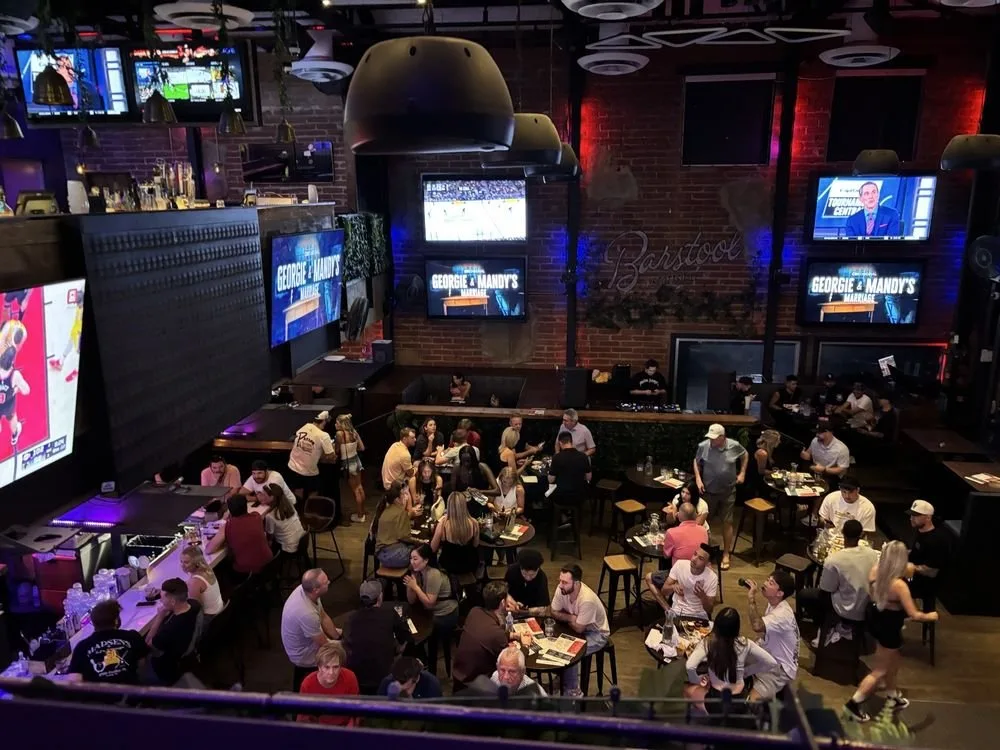 A lively sports bar with multiple television screens showing sports games, patrons sitting and standing around tables, servers attending to customers, and a brick wall with lighting that creates a vibrant atmosphere.