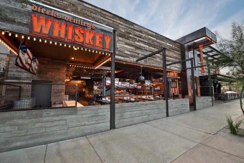 Outdoor view of a bar or restaurant with a sign that reads 'Dierks Bentley's Whiskey Row' and American flags, featuring a modern rustic design with wood and metal elements.