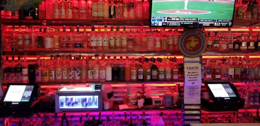Shelves stocked with various bottles of alcohol, illuminated with pink and red lighting, with a television screen displaying a baseball game at the top.