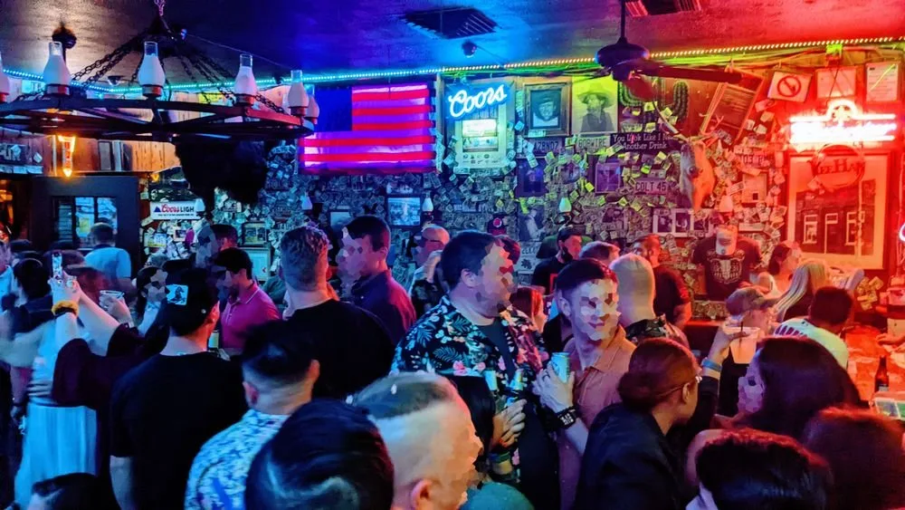 Crowded bar or nightclub with neon signs, including Coors and neon American flag, and dim colorful lighting. People socializing, drinking, and taking photos.