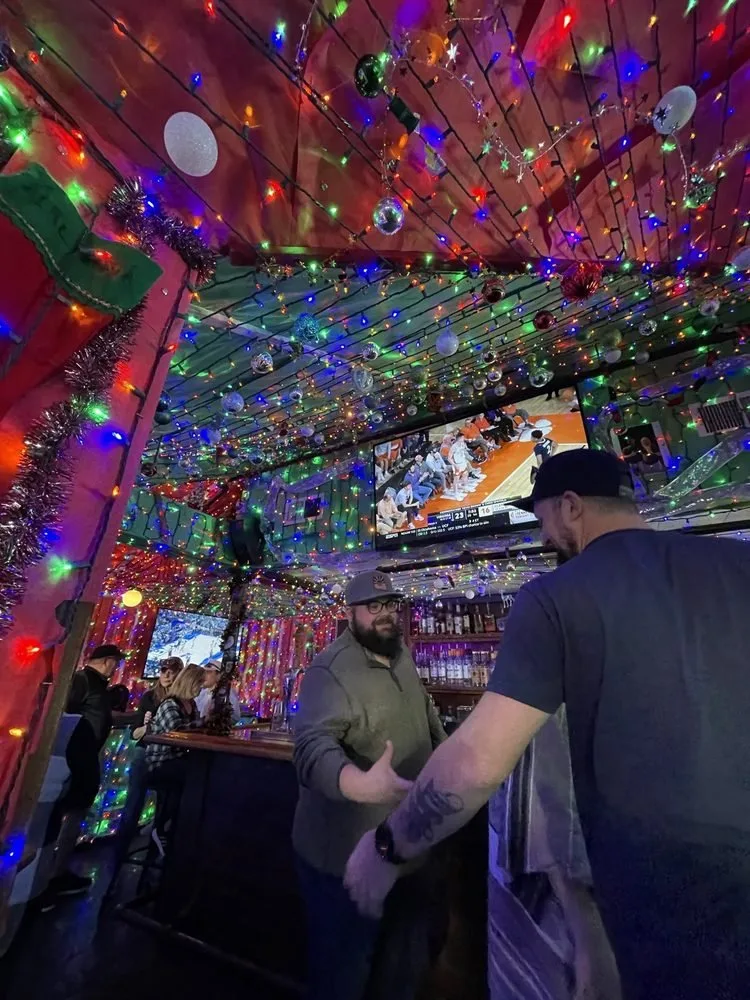 People in a bar decorated with colorful string lights, ornaments, and tinsel for Christmas, with a large TV displaying a sports game.